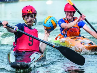 University of Southampton students playing kayak polo on a river, wearing red helmets, with one student holding a yellow and blue ball.
