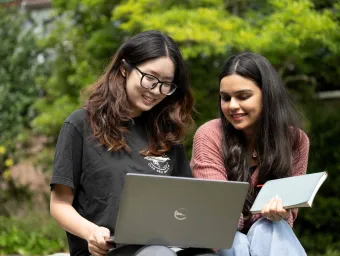 Two students looking at a laptop. They are sitting outside with green shrubs behind them.