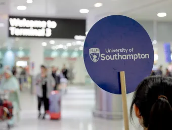 A UoS ambassador waits to greet international students at an airport terminal. She holds a sign bearing our crest and reading "University of Southampton"