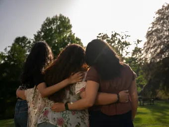 Three international students put their arms around each other and look away from the camera, towards a horizon lined with lush green trees and bright sunny skies