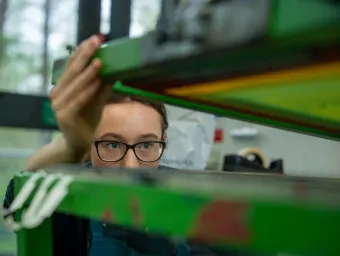 A person wearing glasses adjusts or inspects equipment in a laboratory or workshop setting, viewed through green-tinted panels. The image focuses on close observation and hands-on technical work.