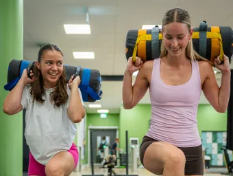 Two students performing weighted walking lunges with sandbags in the university gym.