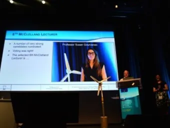 Presentation at the 8th McClelland Lecture showing a speaker on a large screen, with a wind turbine model displayed on stage