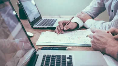 Close up view of two people working at a desk with laptops, paper and pens