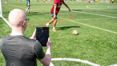 Dr Devon Lewis standing by a football pitch as a player kicks the ball. Devon is holding an iPad that shows live data streamed from a wearable sensor.