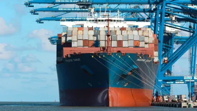 A heavily laden cargo ship sits under cranes by the dockside