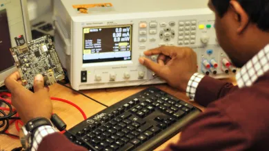 A man in a brown jumper examines a circuit board at a desk