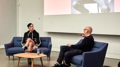 Charles Jeffrey and Professor Jonathan Faiers sit on blue sofas with a small table between them. Jonathan is facing away from the camera and has an arm raised as he speaks. Charles is facing Jonathan and appears to be concentrating on what Jonathan is saying. There is a projector behind them showing a person wearing a pinstripe suit.