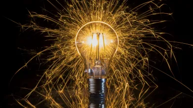 An illuminated light bulb with a vibrant shower of golden sparks radiating around it, resting on a reflective surface with a visible reflection below.