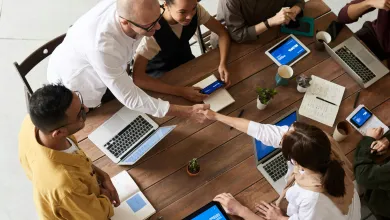 A top-down view of a diverse group of six professionals seated around a wooden table with laptops and tablets, engaged in discussion during a collaborative meeting in a modern workspace.