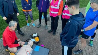 A group of people is observing a CPR training demonstration outdoors, with one person performing the demonstration on a training mannequin next to an AED device, in a sunny, grassy area.