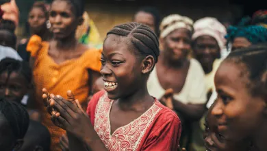 A young girl with braided hair claps and smiles amidst a group of villagers outdoors, capturing the vibrant spirit of Sierra Leone.
