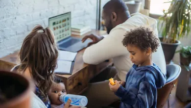 Family at home with father working on laptop, mother feeding baby, and young child playing nearby.