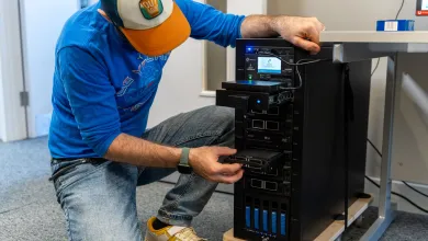 A person is inserting or removing a hard drive from a multi-bay server or data storage system in a tech workspace. They are wearing a blue shirt, jeans, and a cap with the label "Outdoor."