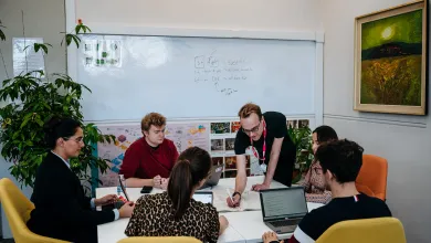A group of people is collaborating around a table in a modern office space, with laptops, papers, and a whiteboard displaying diagrams and notes. One person is leaning over, writing in a book, while others focus on their discussions and screens.