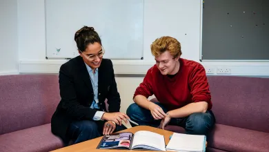 Two people are sitting on a couch in an office space, reviewing a document or booklet together. The woman is smiling and pointing at the material, while the man attentively listens and looks on.