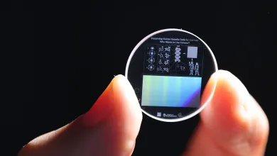 Zoomed in view of a hand holding a 5D memory crystal between two fingers. The memory crystal is a small clear disc with diagrams of human DNA on the surface.