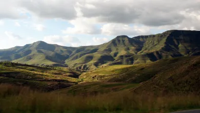 A landscape of rolling hills with long grass in the Lesotho Highlands. There is a mountain range on the horizon, and the sky is mainly cloudy with some clear blue areas.