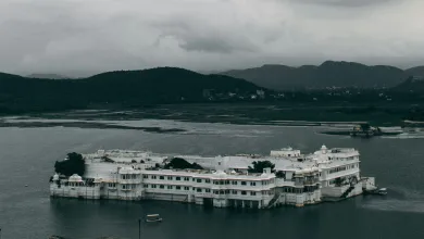 The Taj Lake Palace in Udaipur, Rajasthan, India. It is a rainy day and there are dark clouds surrounding the hills in the distance.