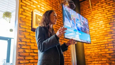 A woman is photographed from below, shown speaking in front of a digital screen.