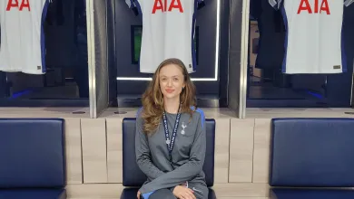 Emma Green wearing a Tottenham Hotspur tracksuit, is sitting on a chair with 3 Tottenham Hotspur shirts hanging behind her.
