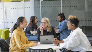 Three people sat a desk talking. The person in middle has a laptop open on the desk.