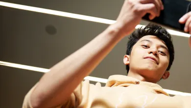 A student looking down at the camera while using their smartphone, which they are holding horizontally. The student is wearing a pale yellow hoodie, and stood in a corridor which is well-lit by several fluorescent lighting strips.