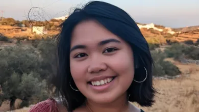 Amatta Mirandari smiling at the camera. She is wearing a dark red muslin top. There is scrubland and blue sky in the background.