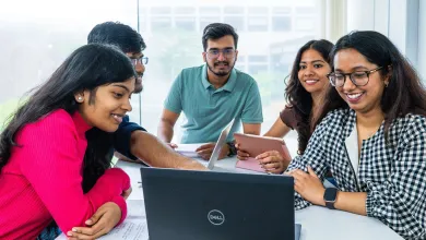 A group of university students work together at a table, smiling and using laptops and tablets.