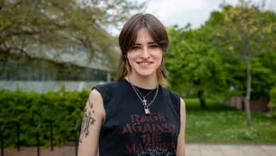 A student stands smiling outside the Physics building dressed casually in a sleeveless band T-shirt.