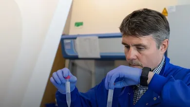 Professor Mark Cragg working in the lab. He is wearing a blue lab coat and holding two pipettes, one is each hand.