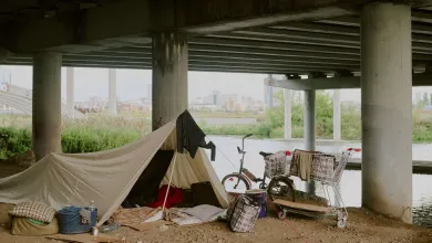 A makeshift shelter set up under a bridge near a river, with personal belongings, a bicycle, and a shopping trolley. The image reflects urban homelessness and temporary living conditions