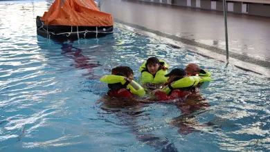 Four people wearing bright yellow life jackets and red immersion suits are floating in a swimming pool, huddled together in a survival position. 