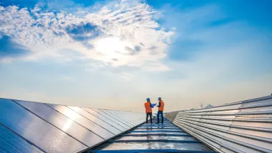Two technicians in protective gear on a rooftop with solar panels under a bright, cloudy sky.