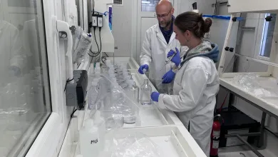 Two people in a laboratory preparing water samples, wearing lab coats and gloves, and handling bottles and equipment on a long workbench.