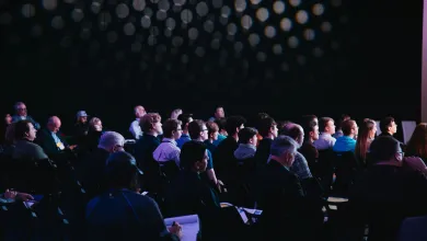A three quarter view from behind of conference attendees sat in rows. The room is mostly dark and there is light coming from the front of the room, out of the shot. 