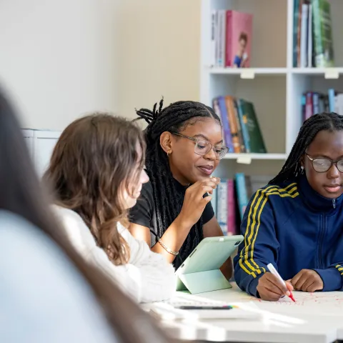 Psychology students sitting around a table. They are writing and mapping out work on a large piece of paper on the table. There are book shelves in the background.