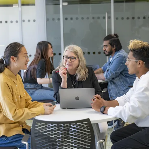 Three people sat a desk talking. The person in middle has a laptop open on the desk.