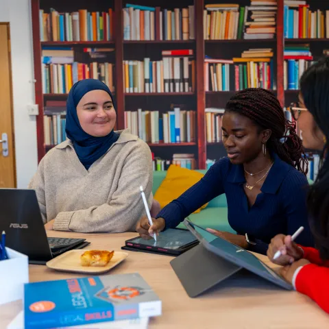 Nora and 2 friends study together in a library. They have their laptops open on the table in front of them.