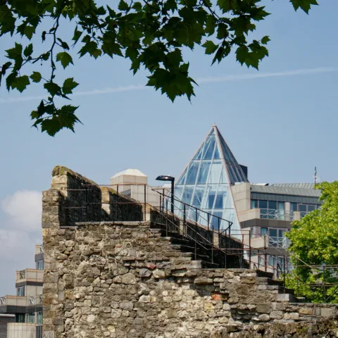 View of a historic stone wall with staircase and metal railings in the foreground, juxtaposed against a modern building featuring a glass pyramid roof. The scene includes leafy tree branches above and a traditional street lamp on the right, highlighting the contrast between medieval and contemporary architecture in an urban setting.