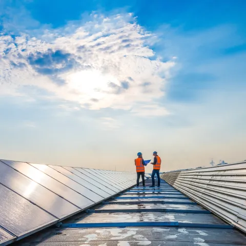 Two technicians in protective gear on a rooftop with solar panels under a bright, cloudy sky.