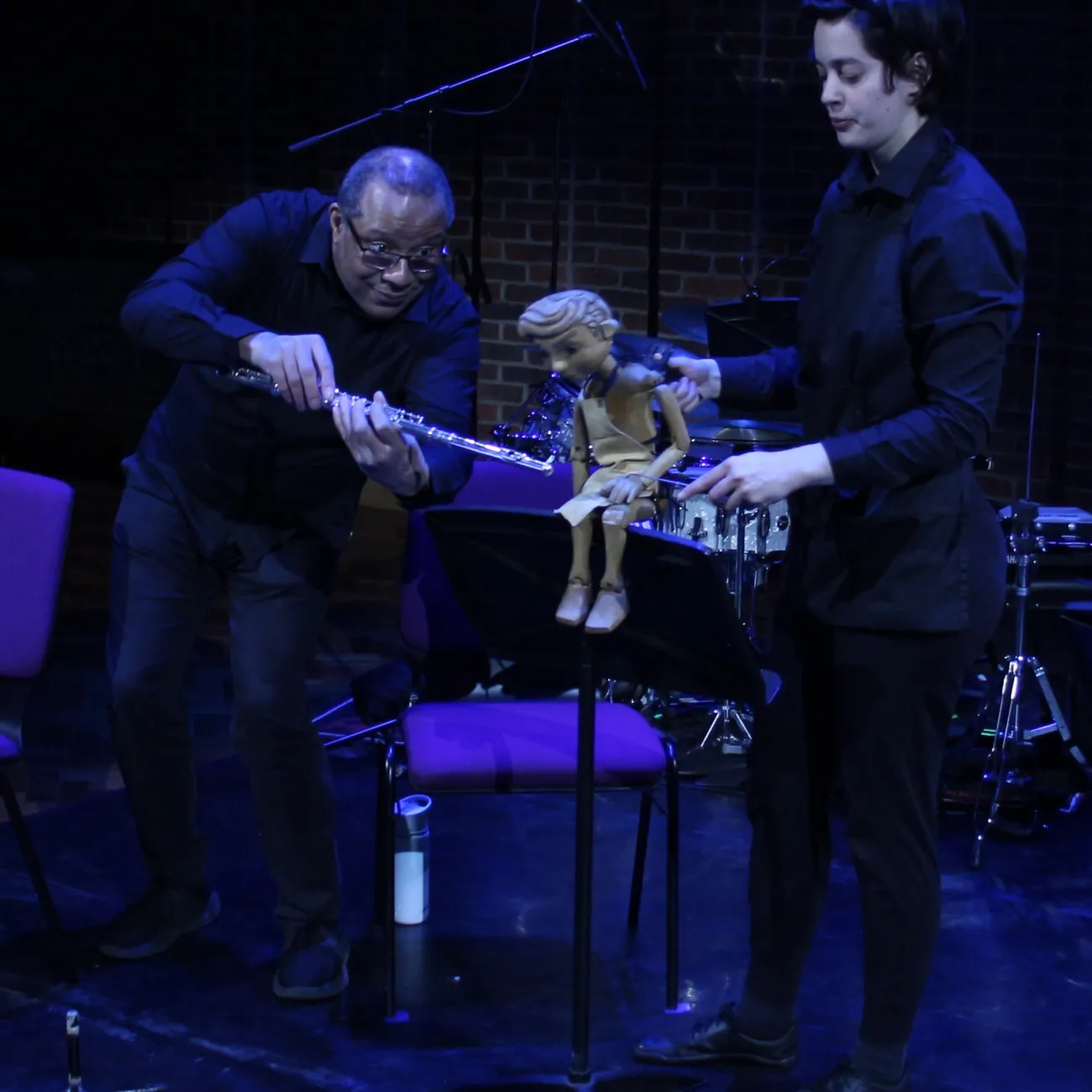 A wooden puppet is positioned on a music stand while one performer holds it and another adjusts a flute nearby. Musical instruments, including drums and chairs, are visible in the background under stage lighting.