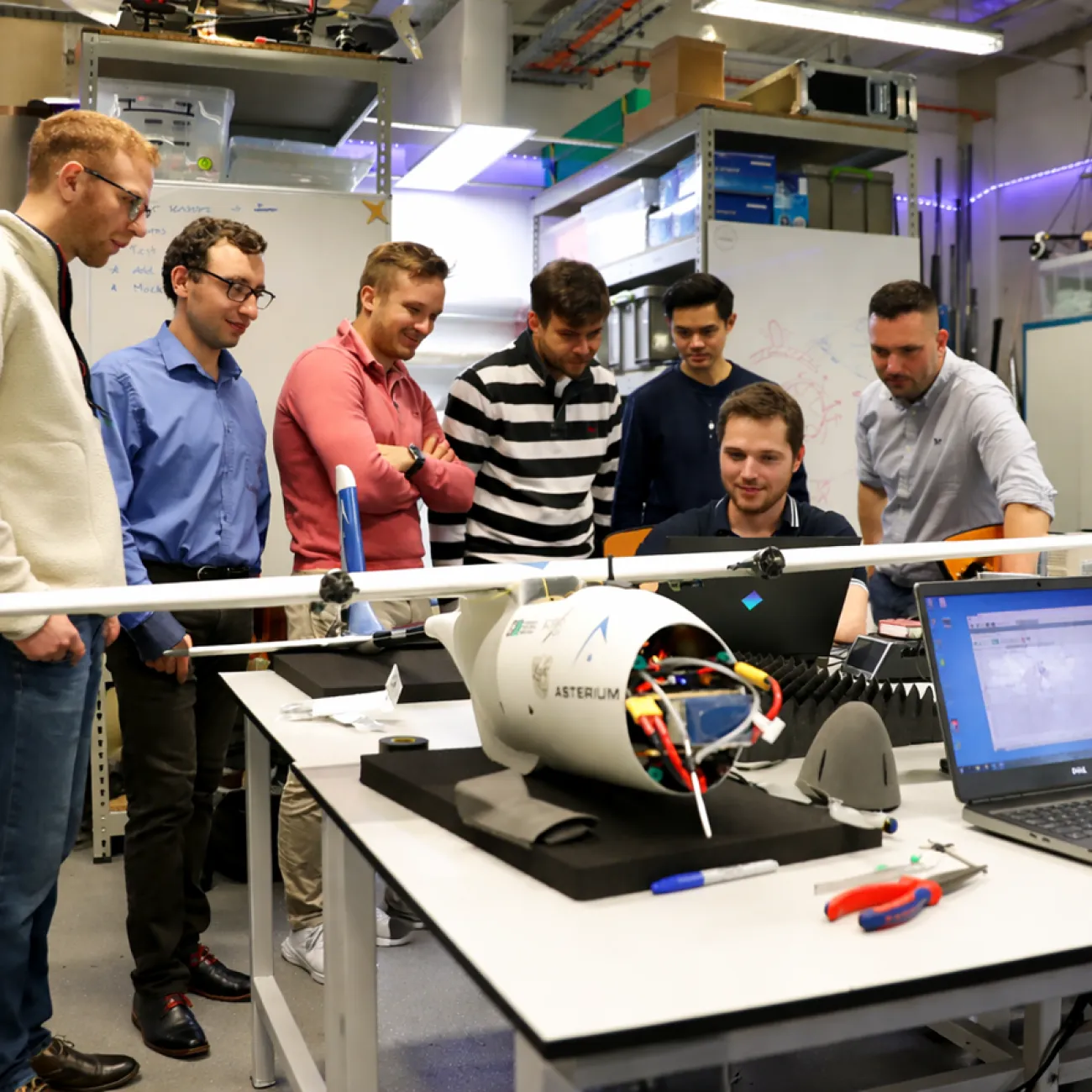 A group of university researchers gathers around a Uncrewed Aerial Vehicle on a workbench in a lab.