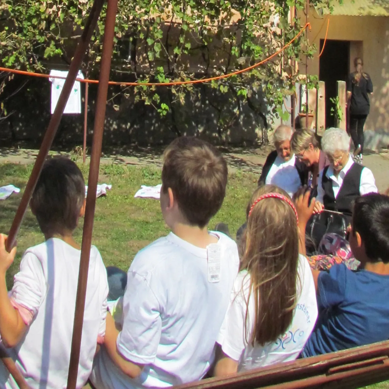 Several children sit on a swing in a green space. Two Romanian women and an interviewer are seated together on a bench in the background, talking.