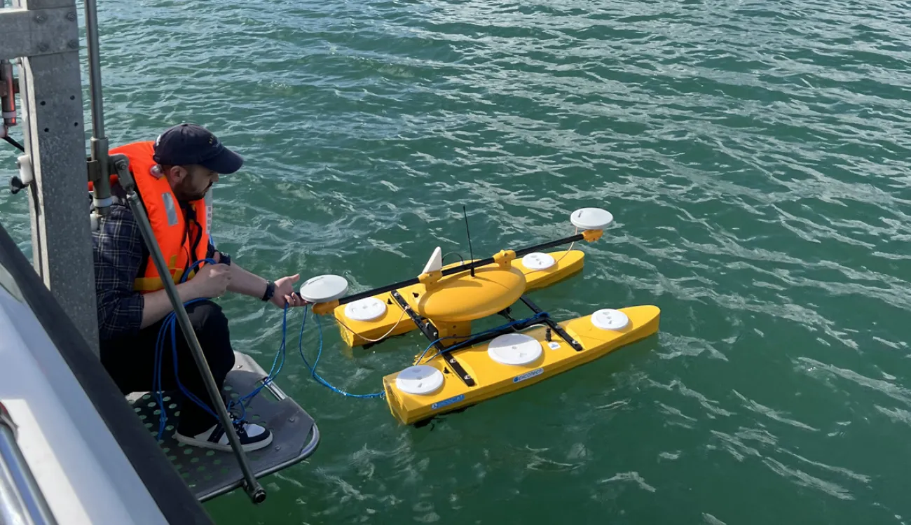 A researcher wearing an orange life jacket deploys a bright yellow, autonomous water surface vehicle to map seabed habitats.