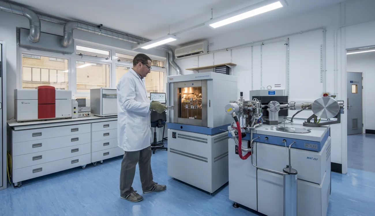A researcher wearing a white lab coat, and gloves, uses large advanced geochemistry facilities.