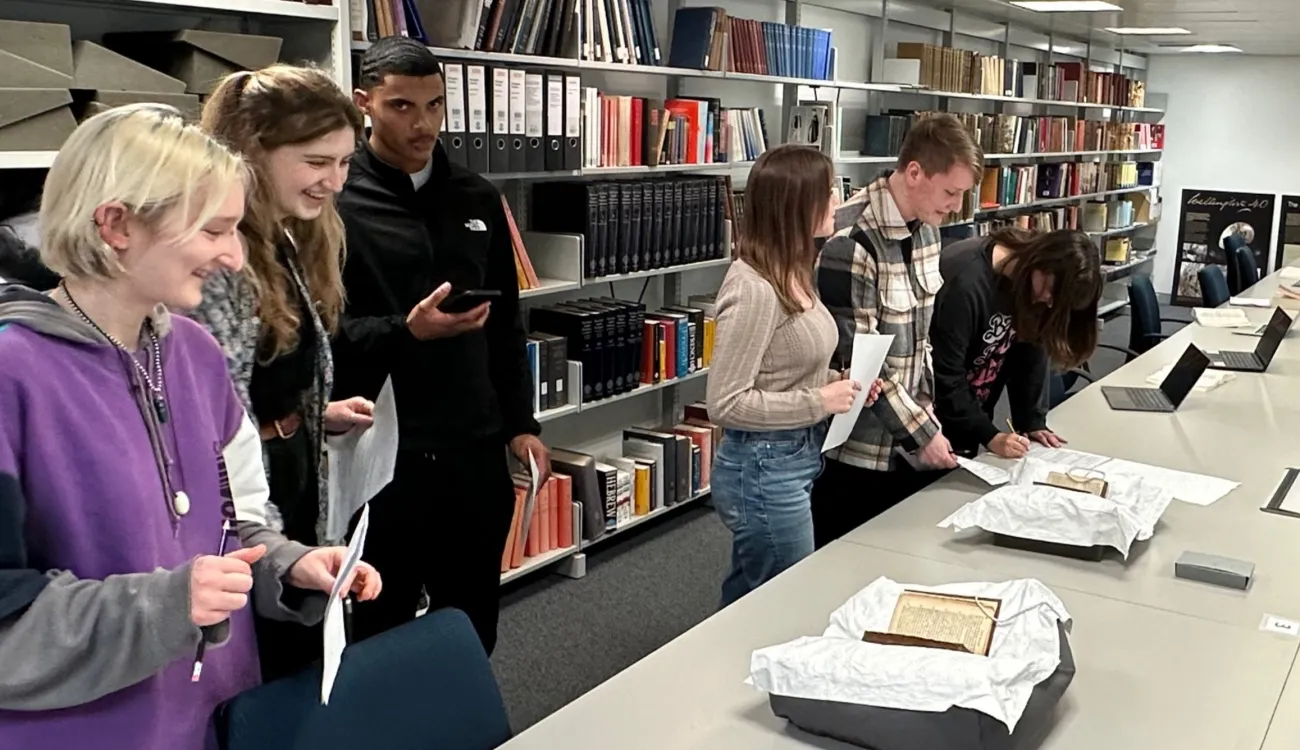 Two groups of three students are standing beside a long table. They are looking at books that are visibly old, with yellowing pages. The books sit open on cushions topped with white cloths. The students are holding pens and paper and one is making notes. Behind them, shelves of books and folders stretch across the entire length of the wall. 