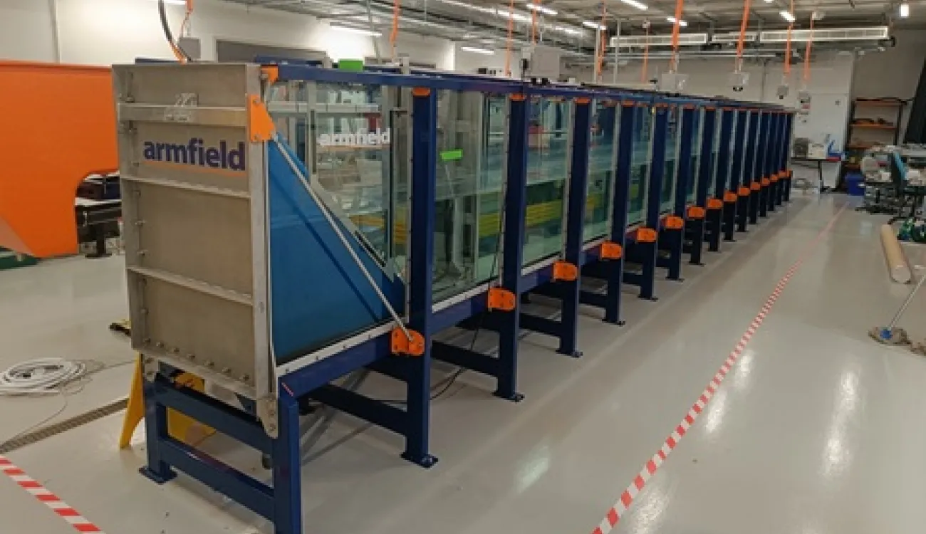 Long glass-sided wave tank in a laboratory, supported by a blue metal frame with orange fittings, and an Armfield wave generator positioned at one end.