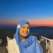 Woman wearing a blue hijab with a flower, smiling at sunset by the sea.