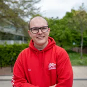 A headshot of a student on a university campus. They are wearing a red hoodie and glasses.
